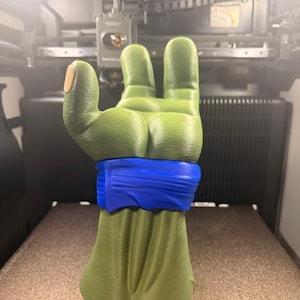 K&ouml;nnte beinhalten: Eine 3D-gedruckte Skulptur einer gr&uuml;nen Hand mit einem blauen Armband. Der Daumen hat einen hellbraunen Nagel. Die Hand zeigt eine Geste, bei der drei Finger hochgehalten werden. Die Skulptur befindet sich vor einem verschwommenen Hintergrund eines 3D-Druckers.