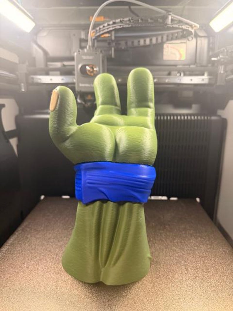 K&ouml;nnte beinhalten: Eine 3D-gedruckte Skulptur einer gr&uuml;nen Hand mit einem blauen Armband. Der Daumen hat einen hellbraunen Nagel. Die Hand zeigt eine Geste, bei der drei Finger hochgehalten werden. Die Skulptur befindet sich vor einem verschwommenen Hintergrund eines 3D-Druckers.