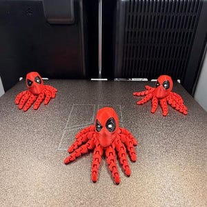 May include: Three red octopus figures with Deadpool masks are displayed on a gray surface. The figures are 3D-printed and have articulated tentacles. The background includes a 3D printer.