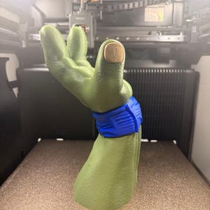 May include: A green, 3D-printed hand with a blue wristband and a tan-colored nail. The hand is posed in a gesture, with the fingers slightly curved. The background shows a 3D printer.