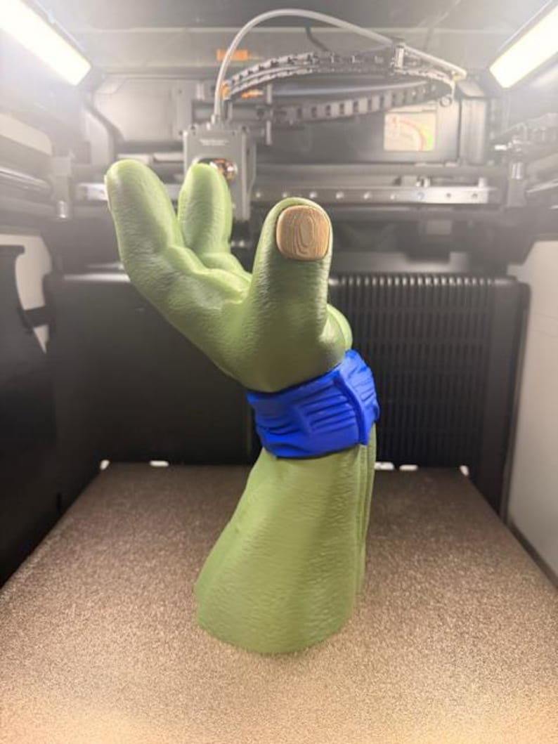 K&ouml;nnte beinhalten: Eine gr&uuml;ne, 3D-gedruckte Hand mit einem blauen Armband und einem hellbraunen Nagel. Die Hand ist in einer Geste gehalten, wobei die Finger leicht gekr&uuml;mmt sind. Der Hintergrund zeigt einen 3D-Drucker.