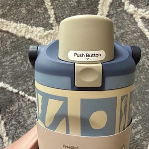 May include: A reusable water bottle with a beige and blue design. The bottle has a push button on the lid and the text "Push Button" is visible. The bottle is cylindrical and has a beige and blue pattern around the body.