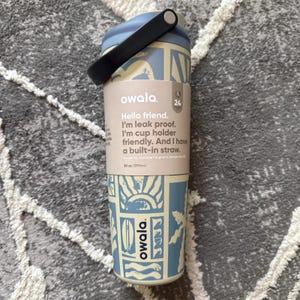 May include: A light blue and cream-colored Owala water bottle with a black handle. The bottle has a patterned design with the text "Hello friend. I'm leak proof. I'm cup holder friendly. And I have a built-in straw." It holds 30 oz.