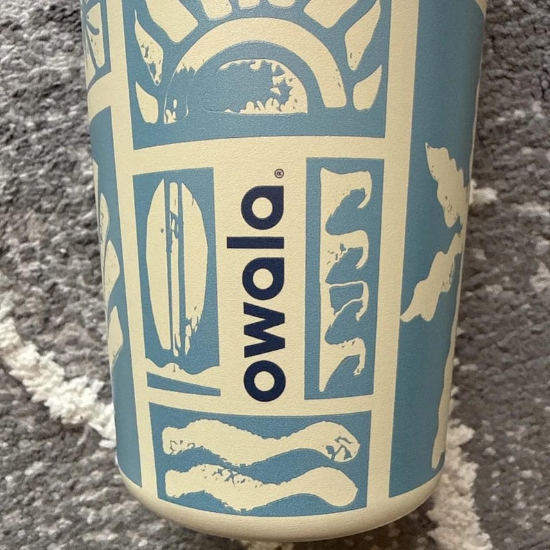 May include: A cylindrical Owala water bottle with a light blue and cream-colored design. The bottle features a repeating pattern of suns, waves, and surfboards. The Owala logo is printed vertically in dark blue.