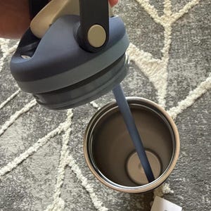 May include: A stainless steel tumbler with a dark blue lid and a matching straw. The lid has a handle and a light beige button. The interior of the tumbler is visible, showing a light-colored base.