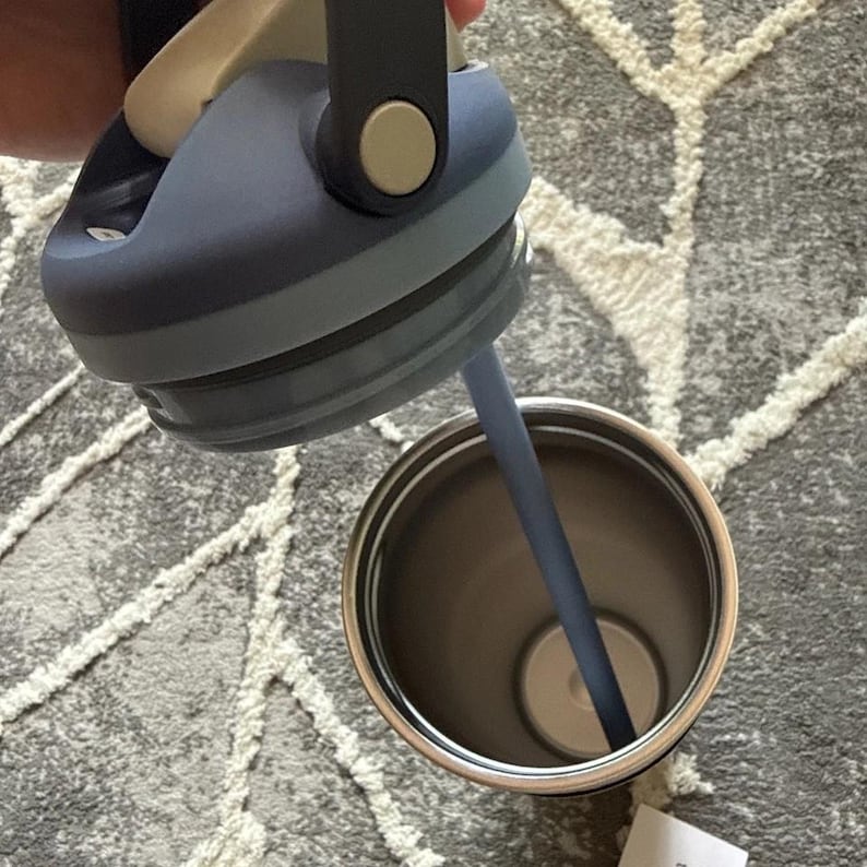 May include: A stainless steel tumbler with a dark blue lid and a matching straw. The lid has a handle and a light beige button. The interior of the tumbler is visible, showing a light-colored base.