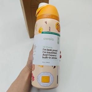 Może przedstawiać: Beżowa butelka na wodę Owala z pomarańczową pokrywką i akcentami. Butelka ma kwiatowy i spożywczy wz&oacute;r z napisem "Hi there. I'm leak proof. I'm insulated. And I have a built-in straw." Mieści 945 ml.