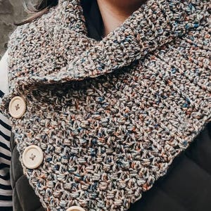 Può includere: Uno scaldacollo in maglia grossa realizzato con un filato variegato nei toni del marrone, grigio e blu. La sciarpa presenta un design a colletto ripiegato, fissato su un lato da tre grandi bottoni rotondi in legno di colore chiaro.