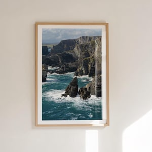 Puede incluir: Una impresión enmarcada de un paisaje costero con acantilados dramáticos y el océano. La imagen muestra formaciones rocosas oscuras, agua turquesa y un cielo nublado. La obra de arte está encerrada en un marco de madera clara, agregando un toque natural.