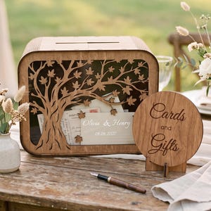Puede incluir: Una caja rectangular de madera con un diseño de árbol cortado con láser en el panel frontal, que contiene sobres con el texto Olivia & Henry 08.08.2027. Al lado hay un letrero circular de madera con el texto grabado Cards and Gifts en un soporte.