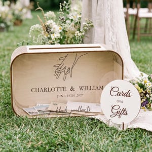 Puede incluir: Una caja rectangular de madera clara para tarjetas con esquinas redondeadas, un dibujo lineal negro de dos manos, el texto "CHARLOTTE & WILLIAM", "JUNE 15TH, 2027" y "joined hands joined hearts". Un letrero redondo blanco dice "Cards and Gifts".