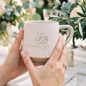 Puede incluir: Una taza de cerámica de color blanco roto con motas y base marrón, que presenta un gran monograma cursivo de las letras ED y el texto ELEANOR & DANIEL 09.02.2025 impreso en color oscuro en la parte frontal, con un asa curva en el lateral.
