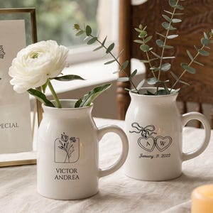 Puede incluir: Dos tazas de cerámica blanca usadas como floreros, cada una con asa y dibujos de líneas negras. Una taza muestra un diseño floral con los nombres VICTOR ANDREA. La otra taza muestra dos relicarios en forma de corazón con las letras A y W y la fecha January 11, 2022.