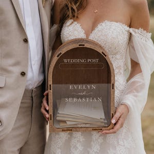 Könnte beinhalten: Eine bogenförmige Hochzeits-Briefbox aus Holz mit einer transparenten Acrylfront. Das Holz trägt die Aufschrift WEDDING POST. Die Acrylplatte ist mit den Namen EVELYN und SEBASTIAN sowie dem Datum 16/05/27 graviert und beschriftet.