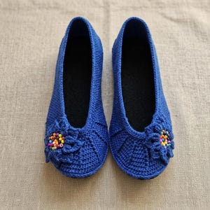 May include: A pair of blue crochet slippers with a floral embellishment on the toe. The slippers have a dark interior and are displayed on a neutral-colored surface. The floral design features colorful beads.