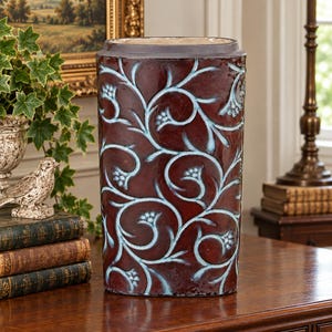May include: A cylindrical ceramic vase featuring a deep burgundy glaze with a raised, light blue vine and floral pattern that wraps around the exterior. The vase has a smooth, dark rim at the top and a wide, stable base with a glossy finish.