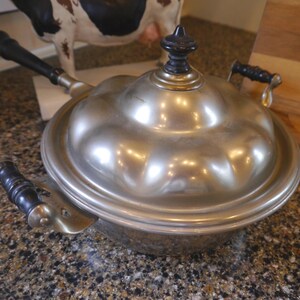May include: A silver-toned, antique metal cooking pot with a domed lid and a black handle. The pot has a scalloped edge and a small, black knob on top. A matching spatula with a black handle rests beside the pot.