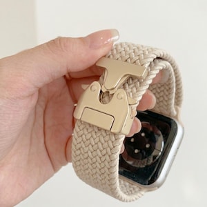 Könnte beinhalten: Beigefarbenes geflochtenes Apple Watch Armband mit goldfarbener Schließe. Das Armband besteht aus einem gewebten Material und verfügt über einen sicheren, einfach zu bedienenden Verschluss. Das Zifferblatt der Uhr ist silberfarben mit einem schwarzen Bildschirm.