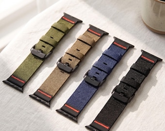 May include: Four nylon watch bands arranged in a row, featuring olive green, tan, navy blue, and black colors. Each band includes a black metal buckle, black hardware connectors, and a distinct red stitched detail at the top and bottom ends.