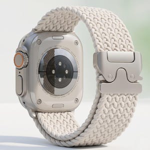 Könnte beinhalten: Eine silberne Smartwatch mit einem cremefarbenen geflochtenen Armband. Die Uhr hat ein orangefarbenes Zifferblatt und einen rechteckigen Bildschirm. Das Armband wird mit einem silbernen Verschluss befestigt. Die Uhr ist für Fitness und Outdoor-Aktivitäten konzipiert.