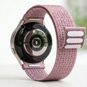 Puede incluir: Primer plano de un reloj inteligente con una correa de tela rosa. La esfera del reloj es negra, con un bisel plateado. La correa se sujeta con velcro. El reloj está etiquetado como "SM-R930 40mm Sapphire Crystal GPS 5ATM".