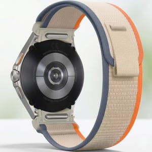 May include: A Samsung Galaxy Watch Ultra smartwatch with a beige, orange, and blue fabric band. The watch has a silver case and a black back with the Samsung logo and other text. The band is secured with a hook and loop fastener.