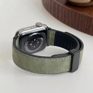 Puede incluir: Un Apple Watch con caja plateada y correa de ante verde salvia. La correa tiene un cierre negro y una parte inferior negra. El reloj se muestra sobre una superficie blanca.