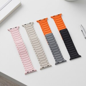 Puede incluir: Cuatro correas de reloj en rosa, beige, naranja y negro. Cada correa tiene un diseño segmentado y está dispuesta sobre una superficie blanca. Las correas están diseñadas para adaptarse a un reloj inteligente.