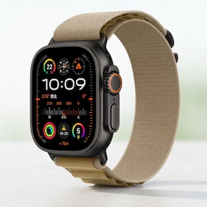 Op de afbeelding: Een zwarte smartwatch met een beige stoffen band. De wijzerplaat toont de tijd 10:09, samen met andere gegevens. Het horloge heeft een oranje accent op de zijkant. De band is gemaakt van een getextureerd, geweven materiaal.