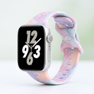 Puede incluir: Un reloj inteligente plateado con una correa colorida y veteada en tonos rosa, morado y azul. La esfera del reloj muestra la hora, con los números 12, 9, 3 y 6 claramente visibles. La correa es de material flexible.