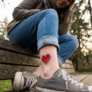 May include: A red heart-shaped patch with a brown border is visible on an ankle. The person is wearing blue jeans and gray sneakers. The background is a wooden bench and a blurred outdoor setting.