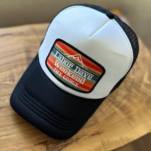 May include: A black and white trucker hat with a white front panel and a black mesh back. The hat features a rectangular patch with the text "Labor Dave Weekend THE GORGE" and a mountain graphic, with a colorful striped design.