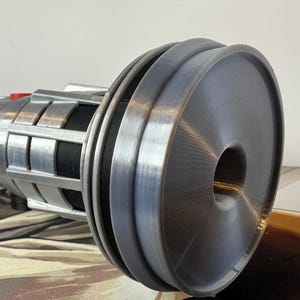 K&ouml;nnte beinhalten: Ein silbernes und schwarzes zylindrisches Objekt mit einem gro&szlig;en, kreisf&ouml;rmigen Ende. Das Objekt hat eine Reihe von Rillen und einen roten Knopf. Das Design umfasst rechteckige und kreisf&ouml;rmige Elemente. Das Objekt liegt auf einer gemusterten Oberfl&auml;che.