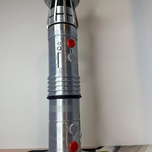 K&ouml;nnte beinhalten: Ein silbernes und graues Lichtschwert-Requisit mit roten und grauen Kn&ouml;pfen. Die Requisite hat ein zylindrisches Design mit gerippten und gerillten Abschnitten. Die Oberseite des Lichtschwertes hat ein detailliertes, geschichtetes Design.