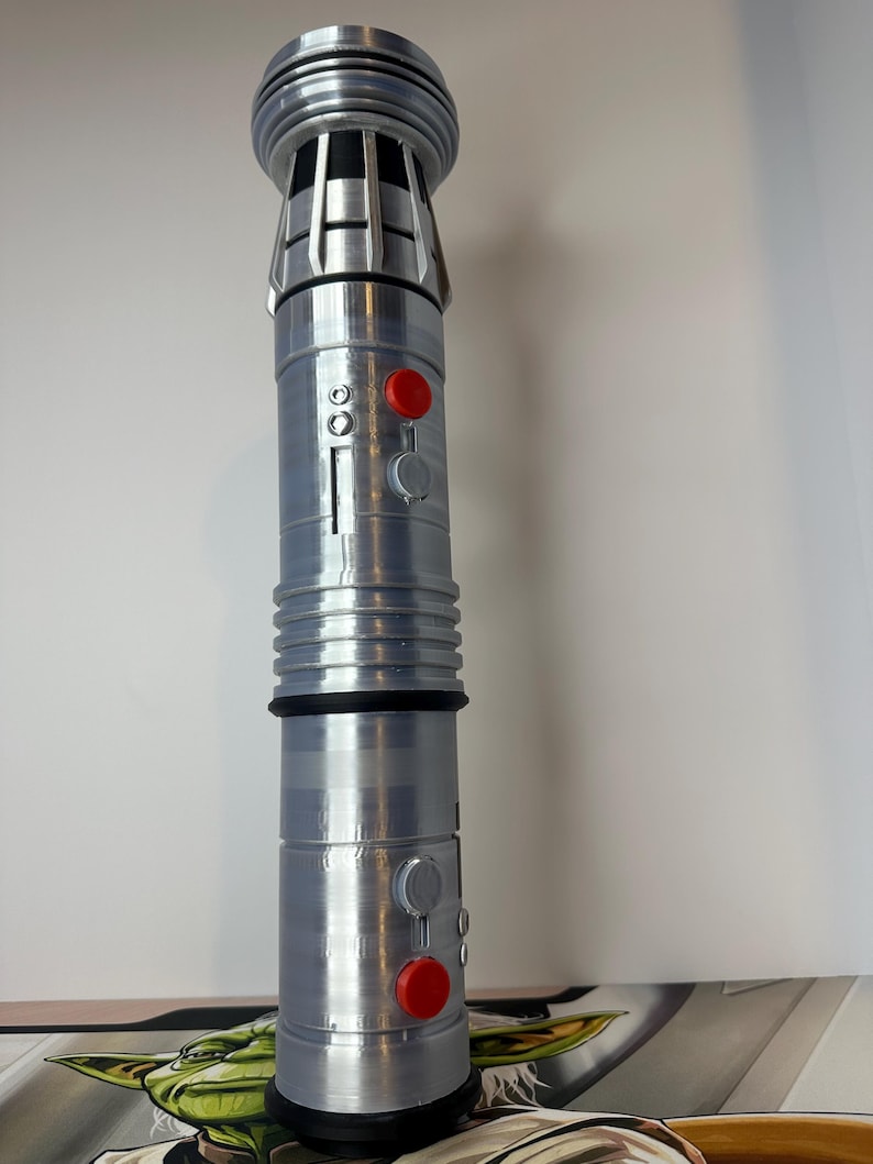 K&ouml;nnte beinhalten: Ein silbernes und graues Lichtschwert-Requisit mit roten und grauen Kn&ouml;pfen. Die Requisite hat ein zylindrisches Design mit gerippten und gerillten Abschnitten. Die Oberseite des Lichtschwertes hat ein detailliertes, geschichtetes Design.