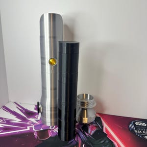 K&ouml;nnte beinhalten: Ein silberner zylindrischer Griff mit einem gelben runden Knopf, eine hohe schwarze zylindrische Komponente und ein kleineres silbernes Metallbasisteil mit einem Gewinde. Die Objekte sind vertikal vor einem wei&szlig;en Hintergrund angeordnet.