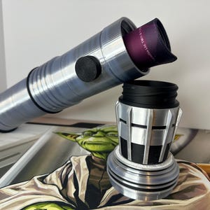 K&ouml;nnte beinhalten: Ein silber-schwarzer Lichtschwertgriff mit einem schwarzen Knopf und einem lila Stoffeinsatz. Der Griff ist zerlegt, wobei der obere Teil zum unteren Teil abgewinkelt ist. Der Hintergrund zeigt eine Yoda-Illustration.