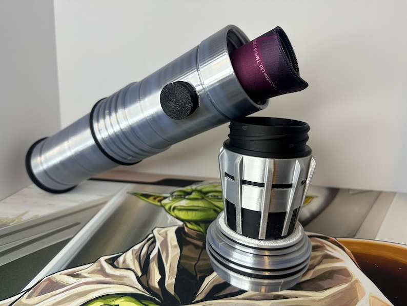 K&ouml;nnte beinhalten: Ein silber-schwarzer Lichtschwertgriff mit einem schwarzen Knopf und einem lila Stoffeinsatz. Der Griff ist zerlegt, wobei der obere Teil zum unteren Teil abgewinkelt ist. Der Hintergrund zeigt eine Yoda-Illustration.