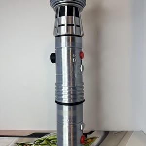K&ouml;nnte beinhalten: Ein silbernes Lichtschwert-Requisit mit detailliertem Design. Der Griff hat schwarze und rote Kn&ouml;pfe und einen geriffelten Griff. Die Oberseite hat ein zylindrisches Design mit einer Reihe von Rippen. Das Lichtschwert ist ein Requisit aus einem Science-Fiction-Franchise.