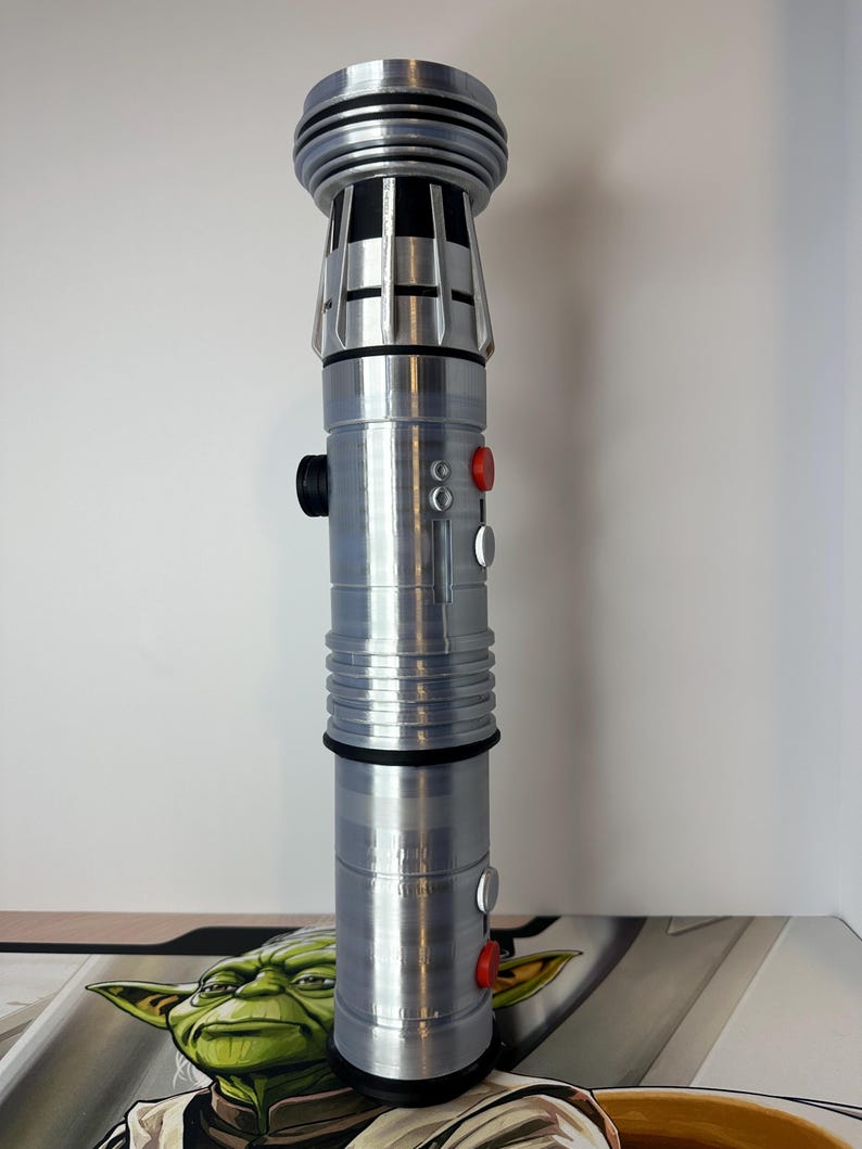K&ouml;nnte beinhalten: Ein silbernes Lichtschwert-Requisit mit detailliertem Design. Der Griff hat schwarze und rote Kn&ouml;pfe und einen geriffelten Griff. Die Oberseite hat ein zylindrisches Design mit einer Reihe von Rippen. Das Lichtschwert ist ein Requisit aus einem Science-Fiction-Franchise.