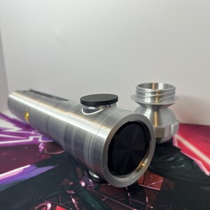 K&ouml;nnte beinhalten: Ein zylindrischer Griff aus silberfarbenem Metall mit einem mattschwarzen runden Knopf an der Seite und einem schwarzen, strukturierten runden Einsatz an der Oberseite. Ein separates silberfarbenes Metallteil mit Gewindehals steht dahinter.