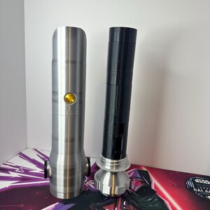 K&ouml;nnte beinhalten: Zwei zylindrische Lichtschwertgriffe stehen aufrecht. Der linke Griff ist silberfarben mit einem runden goldenen Knopf. Der rechte Griff ist schwarz mit einem silbernen Knauf und einem silbernen Emitterteil an der Basis mit geschichtetem Design.