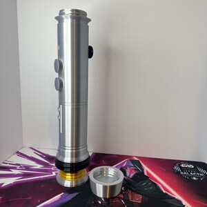May include: A silver-colored cylindrical lightsaber hilt featuring a metallic finish, two circular buttons on the side, a black accent ring, and a gold-colored base component. A separate silver-colored circular cap is positioned next to the hilt.