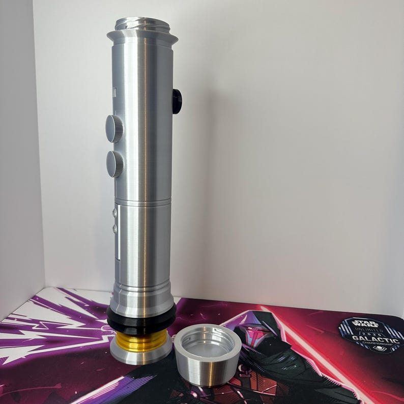 May include: A silver-colored cylindrical lightsaber hilt featuring a metallic finish, two circular buttons on the side, a black accent ring, and a gold-colored base component. A separate silver-colored circular cap is positioned next to the hilt.