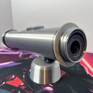 May include: A silver-colored cylindrical tube holder with a flared rim, containing a tightly rolled black textured mat inside. The tube is mounted horizontally on a matching silver-colored conical base, featuring a smooth, matte finish throughout.