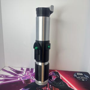 K&ouml;nnte beinhalten: Ein zylindrischer Lichtschwertgriff mit metallisch silbernem Finish an den oberen und mittleren Abschnitten, kontrastiert durch schwarze Komponenten. Er enth&auml;lt zwei kleine gr&uuml;ne runde Kn&ouml;pfe an einem schwarzen Bedienfeld.