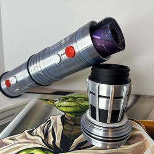 K&ouml;nnte beinhalten: Ein silbernes und schwarzes Schreibtisch-Zubeh&ouml;r-Set im Lichtschwert-Design. Das Lichtschwert ist zylindrisch mit roten Kn&ouml;pfen und einem schwarzen Griff. Die Basis ist ein zylindrischer Becher mit schwarzem Innenraum. Eine aufgerollte Mausunterlage befindet sich im Lichtschwert.