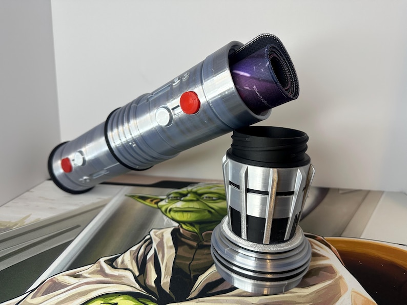 K&ouml;nnte beinhalten: Ein silbernes und schwarzes Schreibtisch-Zubeh&ouml;r-Set im Lichtschwert-Design. Das Lichtschwert ist zylindrisch mit roten Kn&ouml;pfen und einem schwarzen Griff. Die Basis ist ein zylindrischer Becher mit schwarzem Innenraum. Eine aufgerollte Mausunterlage befindet sich im Lichtschwert.