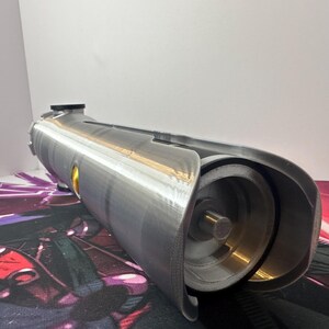 K&ouml;nnte beinhalten: Ein zylindrisches Requisit in Silberoptik mit metallischem Finish, das ein hohles, offenes Ende mit einer inneren kreisf&ouml;rmigen Struktur, einen zentralen Stab und einen kleinen gelben kreisf&ouml;rmigen Akzent an der Seite des Geh&auml;uses aufweist.