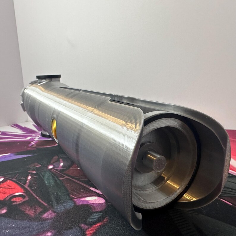 K&ouml;nnte beinhalten: Ein zylindrisches Requisit in Silberoptik mit metallischem Finish, das ein hohles, offenes Ende mit einer inneren kreisf&ouml;rmigen Struktur, einen zentralen Stab und einen kleinen gelben kreisf&ouml;rmigen Akzent an der Seite des Geh&auml;uses aufweist.